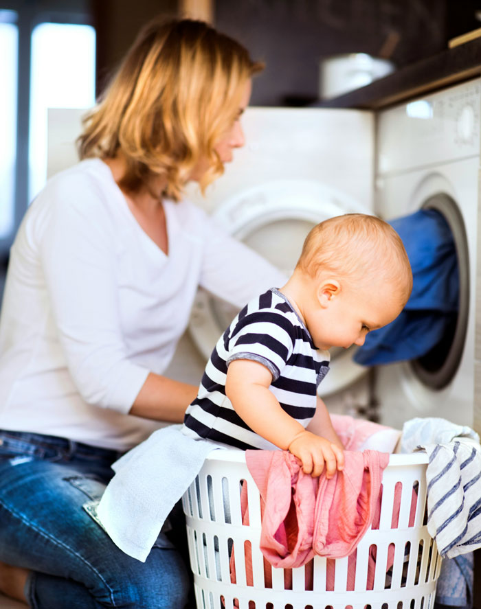 Woman doing laundry with baby sitting in basket, illustrating moments from biggest mistakes described by 68 people.