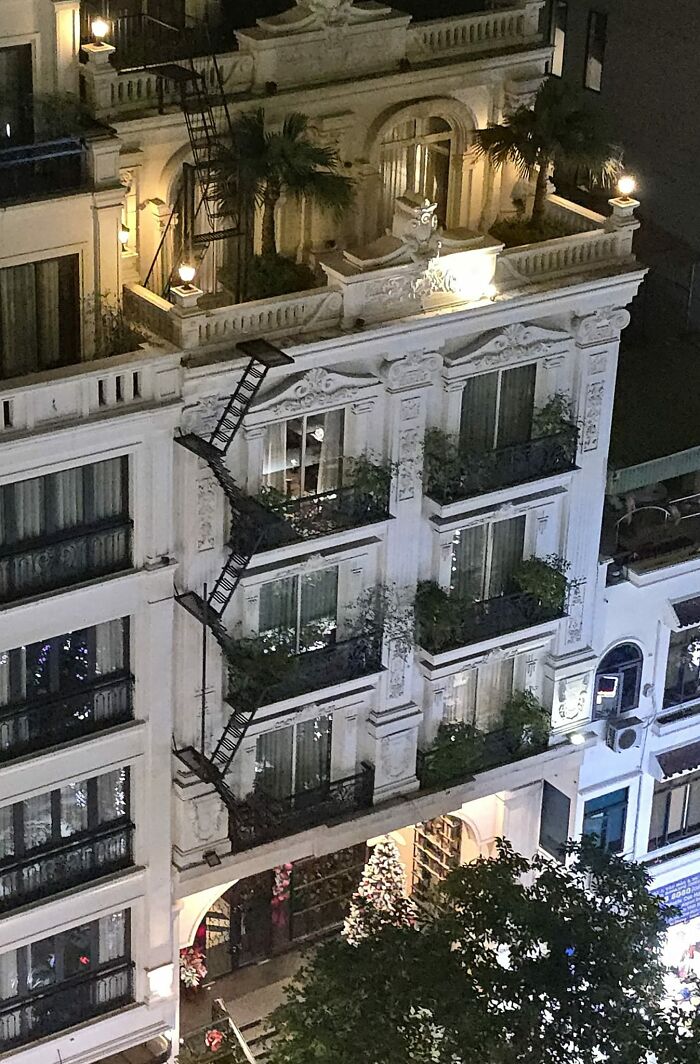 Narrow death stairs attached to a building facade, illuminated at night with plants on balconies and busy street below.