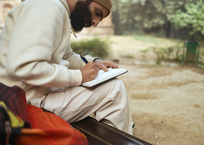 Man with beard wearing beige hoodie and hat writing in notebook outdoors using obsolete things still in daily use