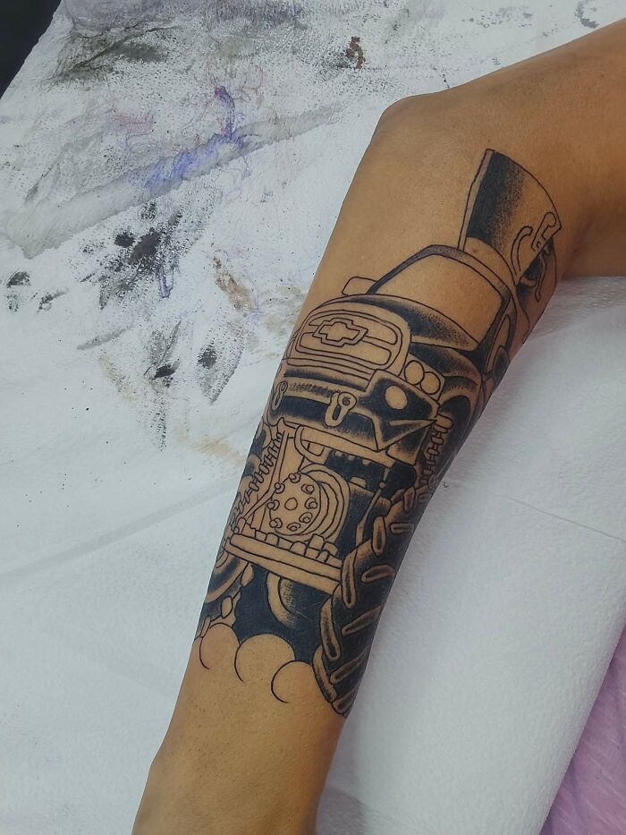 Tattoo of a detailed truck on forearm, showing intricate linework and shading in a bold black ink style.