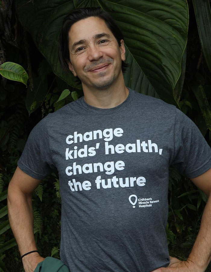 Smiling man wearing a gray t-shirt with a message, standing outdoors among large green leaves, looking way younger.
