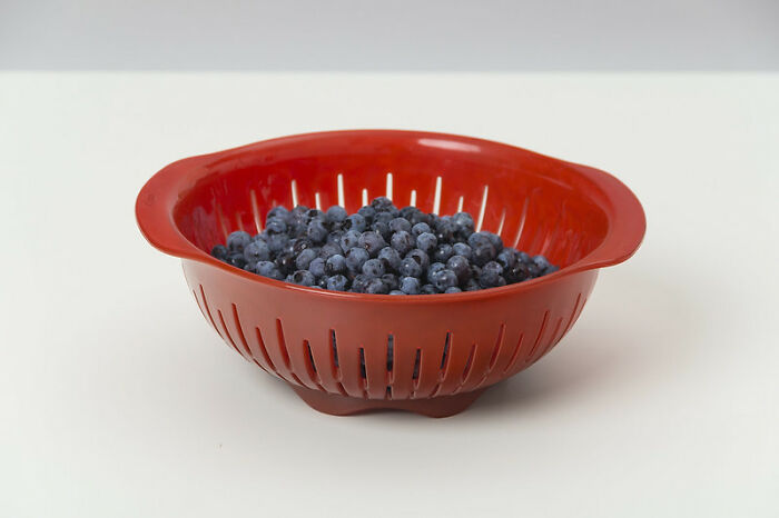 Red colander filled with fresh blueberries on a white surface, illustrating simple and healthy food preparation.