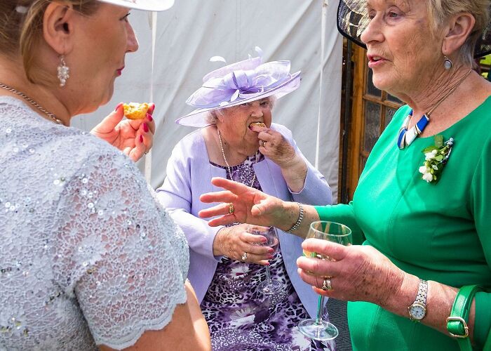 Three elderly women at a wedding reception, dressed in formal attire, enjoying drinks and snacks in a candid moment.