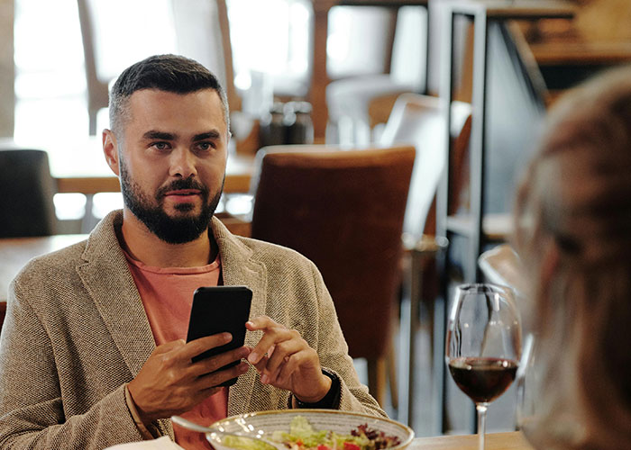 Man on an awkward date looking at his phone across a restaurant table with a glass of wine and salad nearby.