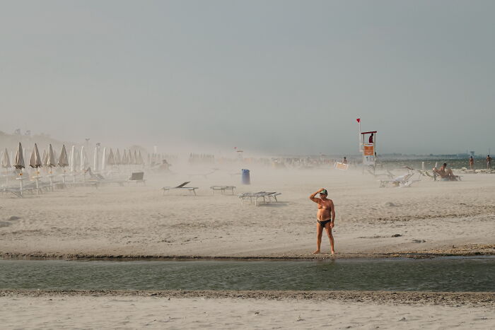 Man standing on a misty beach near empty lounge chairs, captured in a perfectly timed street photo by Luca Regoli.