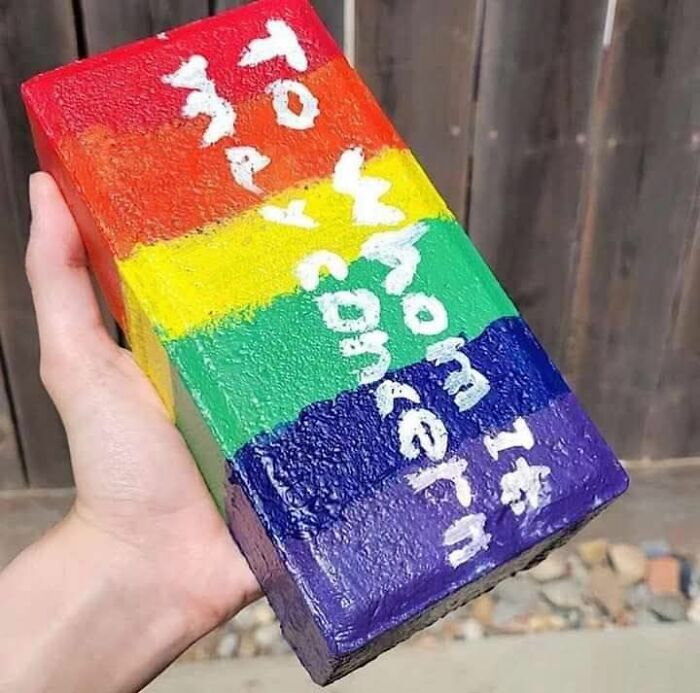 Hand holding a DIY painted brick with rainbow colors and misspelled white text, an example of horrendous craft shaming project.