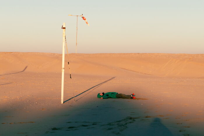 Person lying on desert sand near poles under soft light in a perfectly-timed street photo by Luca Regoli.