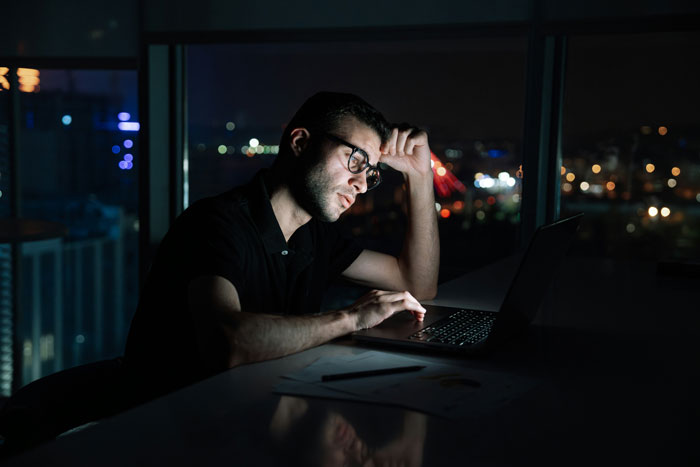 Millennial man in glasses working late on laptop, reflecting deeply, with city lights blurred in the background.