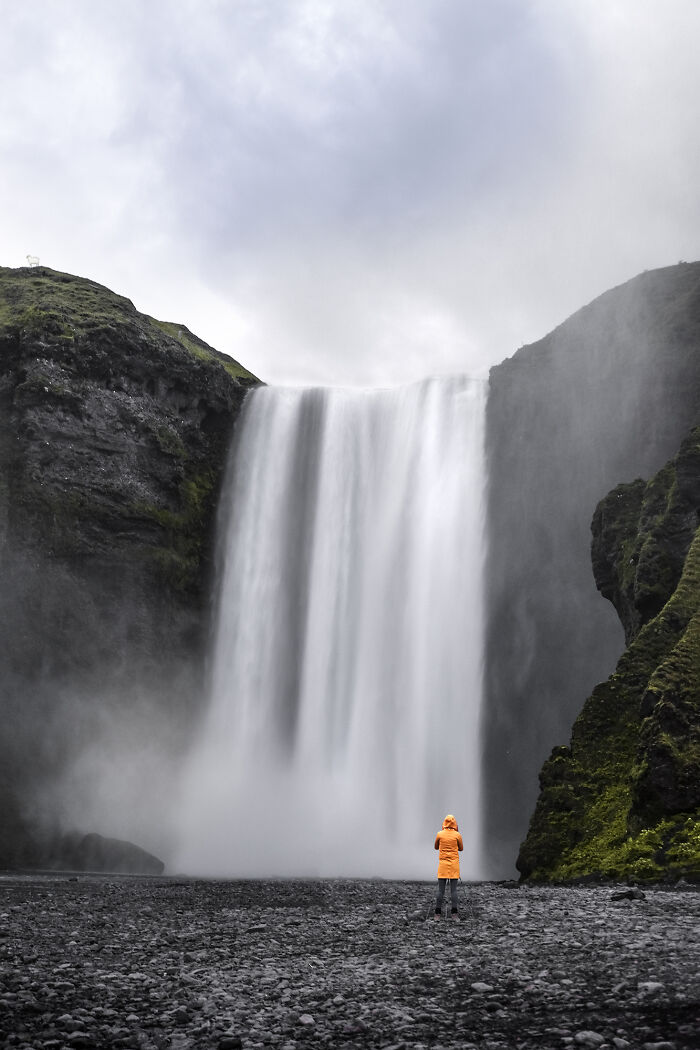 Traveler in orange jacket admiring breathtaking waterfall surrounded by cliffs, showcasing stunning travel photos from Bored Panda community.