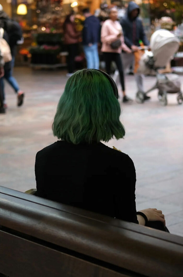 Person with green hair sitting on a bench wearing headphones, captured in a beautiful street shot of everyday life.
