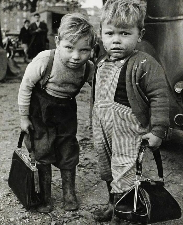 Two young boys in worn clothes holding bags and standing outdoors in a fascinating historical photo from the past.
