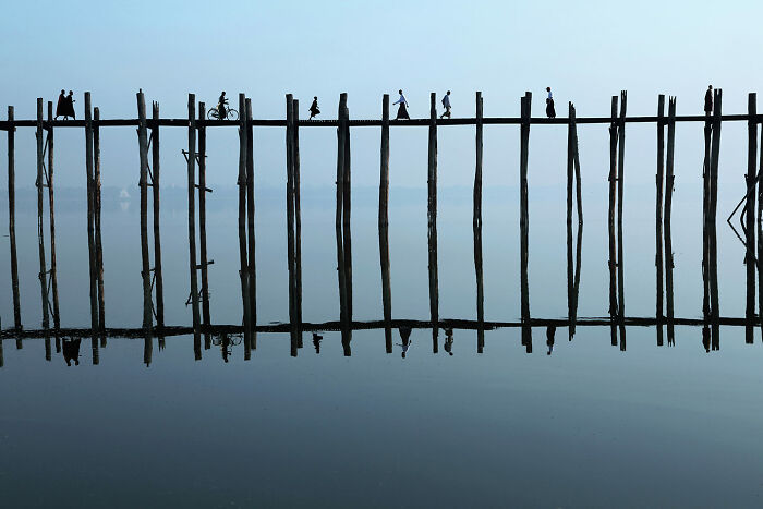 Silhouettes of people walking and cycling on a wooden bridge over calm water in a breathtaking travel photo.