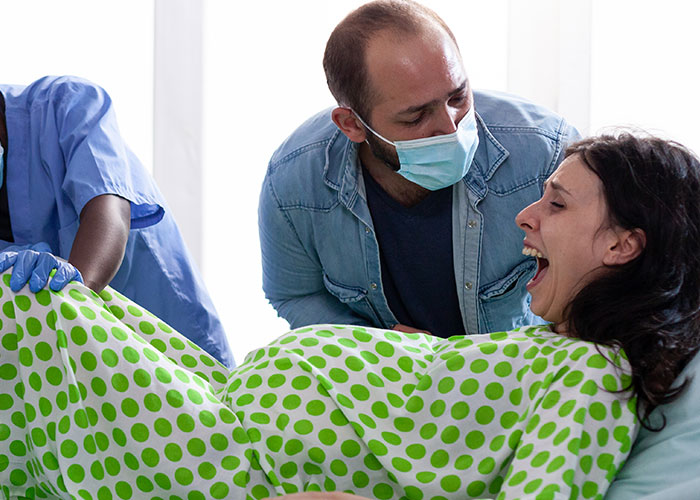 Man wearing mask supports woman in labor inside birthing room as medical staff assist during childbirth experience.