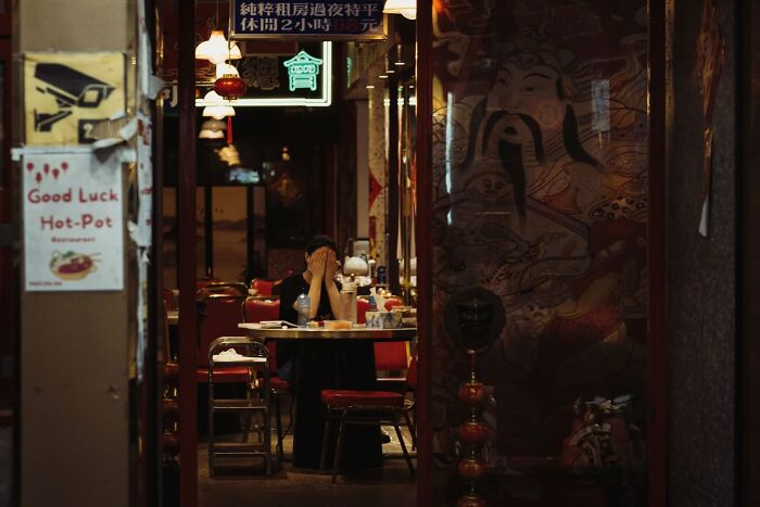 Woman covering face inside dimly lit restaurant captured in candid street photo by Alex McClintock with vibrant urban details.