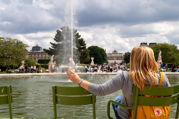 Woman holding a bottle aligned with a fountain jet, a creative and perfectly-timed street photo by Luca Regoli.