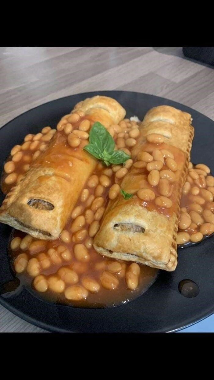 Two baked pastry rolls covered in baked beans and sauce on a black plate, a cursed food picture that might make you say ewww.