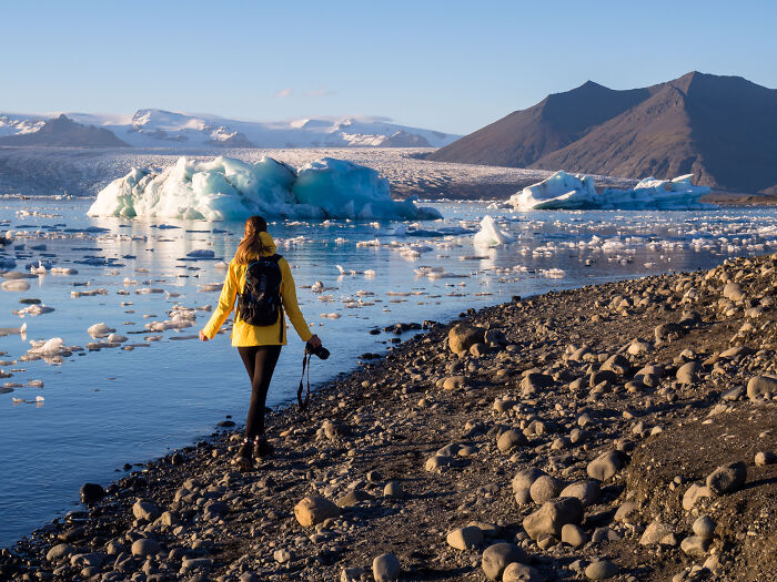 Traveler in yellow jacket walking by icy lake with glaciers and mountains, showcasing breathtaking travel photos from the Bored Panda community.