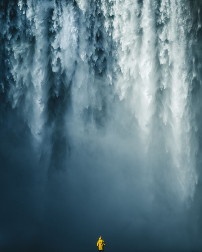 Person in yellow raincoat standing at the base of a massive waterfall in breathtaking travel photos captured by the community.