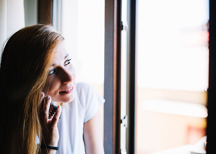 Young woman standing by window smiling and talking on phone, relating to cheating boyfriend and psycho confrontation topic.