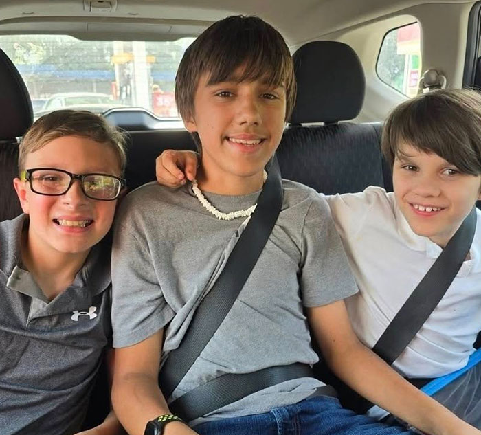 Three teenage boys sitting together in the backseat of a car, smiling and wearing seatbelts.