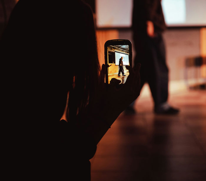 Person capturing a speaker on phone camera in dim light, highlighting toxic dating trends people accept as normal.