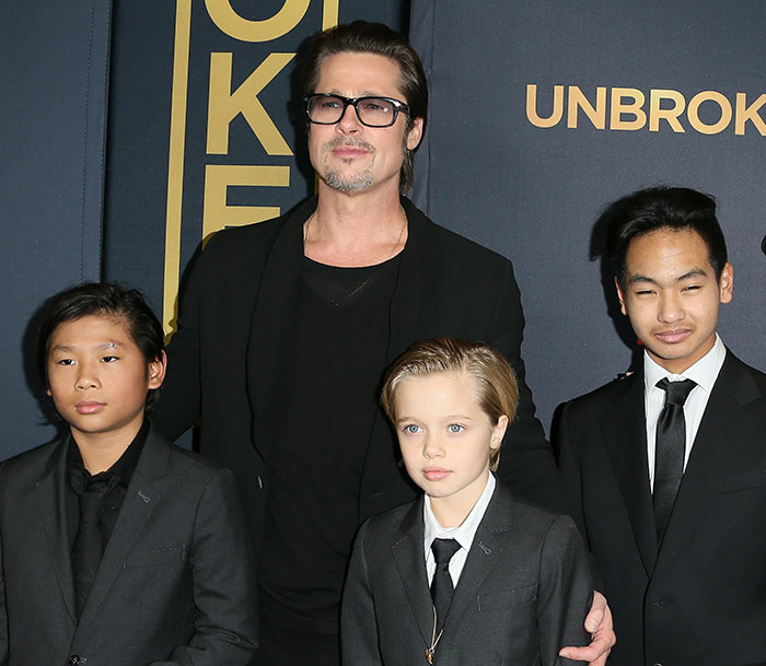 Brad Pitt posing with sons at an event, dressed in black suits and ties against a dark backdrop.