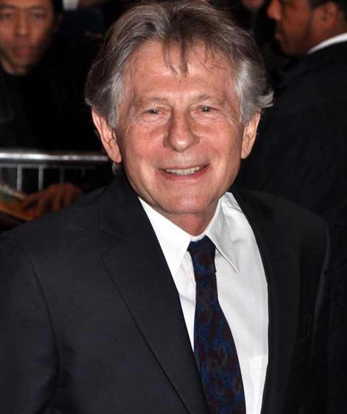 Older man in a black suit and patterned tie smiling at an event related to famous people with past controversies.