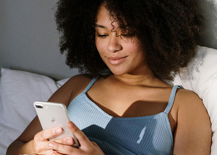 Young woman with curly hair lying on bed, smiling while looking at her phone, relating to wife cheats on husband topic.