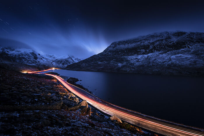 Nighttime travel photo showing light trails on a winding mountain road near a lake, captured by the Bored Panda community.