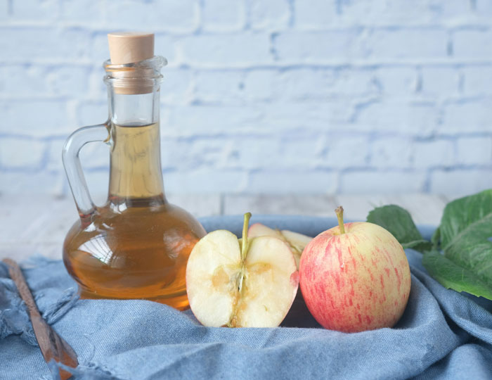 Glass bottle of apple cider vinegar with whole and halved apples on blue cloth, illustrating effective old wives' tales.