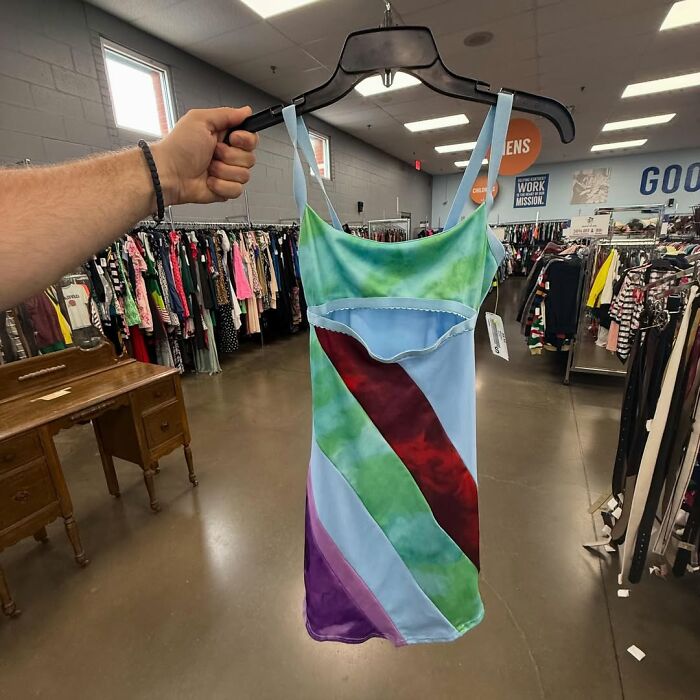 Colorful secondhand dress with blue, green, red, and purple stripes held in a thrift store filled with clothes racks.