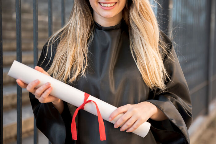 Young woman in graduation gown holding diploma, smiling confidently, illustrating people committed to a lie way too hard.