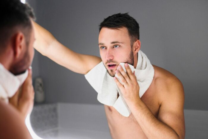 Young man with towel around neck wiping face in bathroom mirror, representing nail biting and grooming habits.