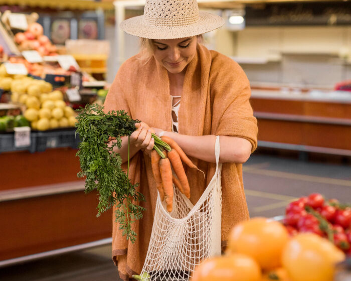 Woman at market putting fresh carrots in reusable bag demonstrating poor people habits despite wealth.