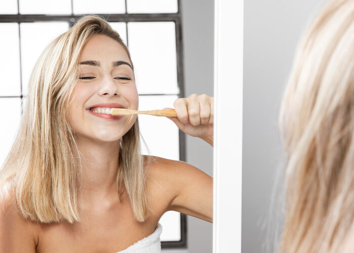 Young woman brushing teeth in bathroom mirror, illustrating nail biting and personal hygiene habits.