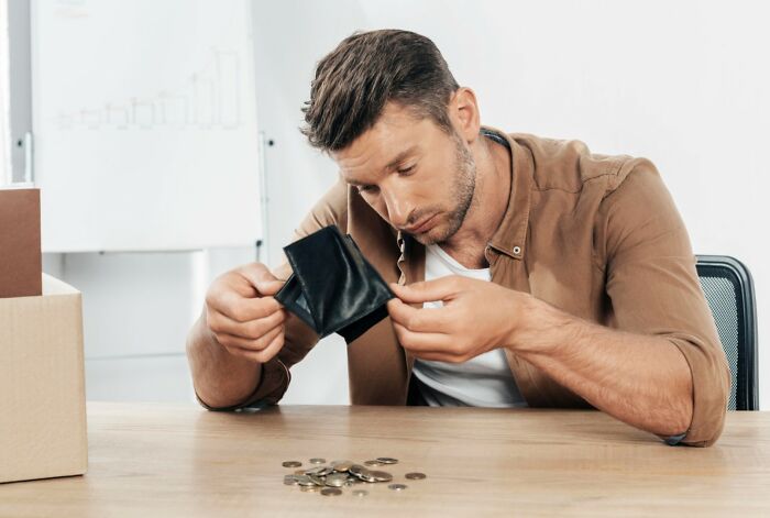 Man committed to a lie way too hard looking disappointed at empty wallet with scattered coins on table indoors.