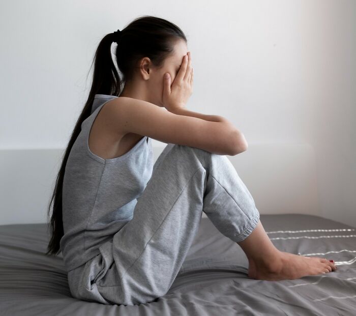Young woman sitting on bed covering her face, showing signs of a difficult childhood and emotional distress in a quiet room.