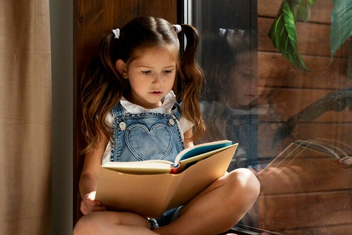 Young girl reading a book by the window, reflecting a calm moment as a good trend slowly disappeared from daily life.
