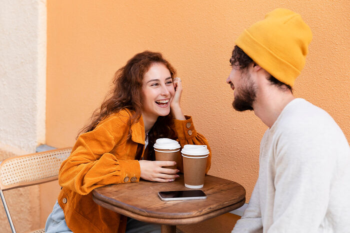 Young couple sharing coffee and laughing during a modern dating meetup at a cozy cafe with warm orange tones.