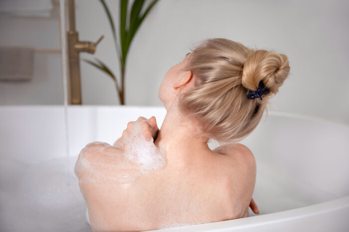 Woman with blonde hair in a bun taking a bubble bath, relaxing while washing her shoulders in a white bathtub.