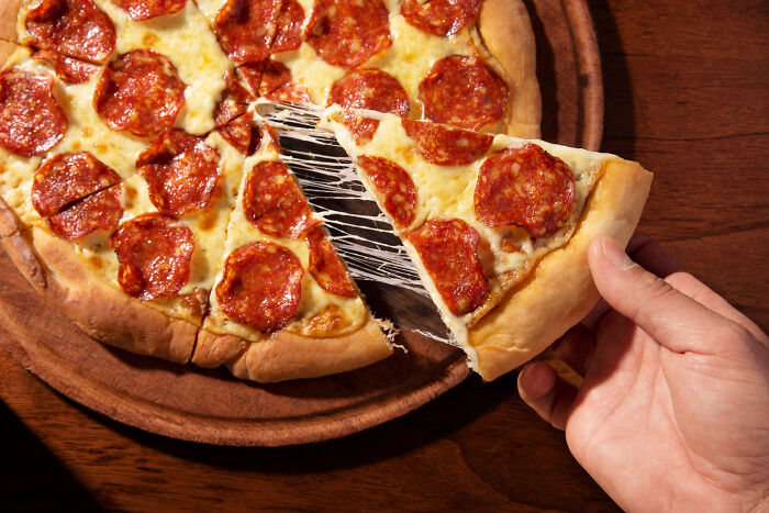 Hand holding a slice of pepperoni pizza with melted cheese being pulled apart on a wooden table.