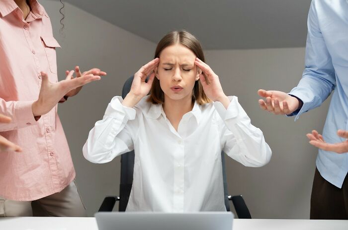 Young woman stressed at work with hands on temples, surrounded by arguing colleagues showing signs of a troubled childhood