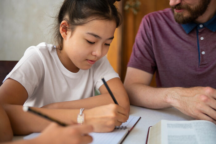 Young girl focused on writing in a notebook with an adult nearby, illustrating signs of a challenging childhood experience.