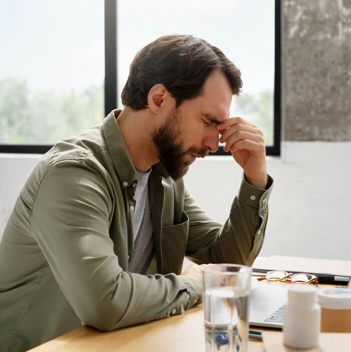 Man sitting at desk with eyes closed and hand on forehead, showing signs of stress related to childhood experiences.