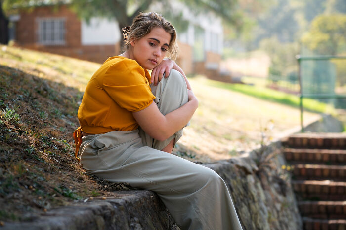 Young woman sitting alone outdoors, showing signs of a difficult childhood through her thoughtful and somber expression.