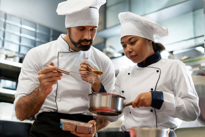 Two professional chefs in white uniforms tasting sauce in a commercial kitchen, showcasing culinary professions. - 10