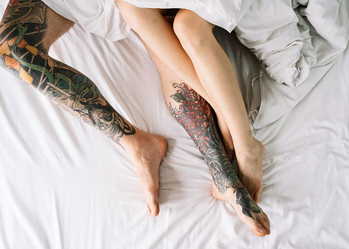 Two people lying in bed showing detailed leg tattoos, representing a take one for the team moment in relationships.