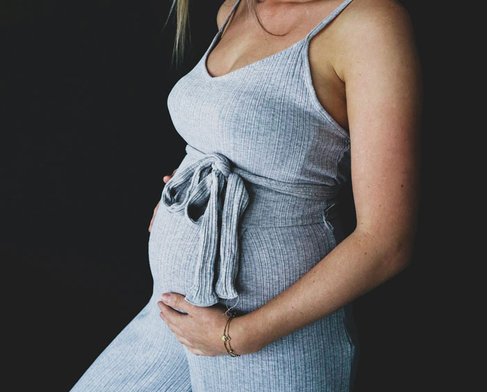 Pregnant woman in a grey sleeveless dress gently holding her belly against a dark background about paying for college.