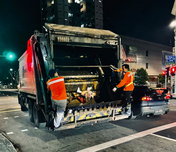 Two sanitation workers in bright orange jackets collecting garbage from a truck at night, highlighting millennials biggest lies.