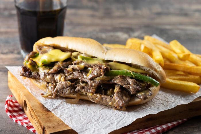 Philly cheesesteak sandwich with melted cheese and peppers served with fries and a soda drink on a wooden board.